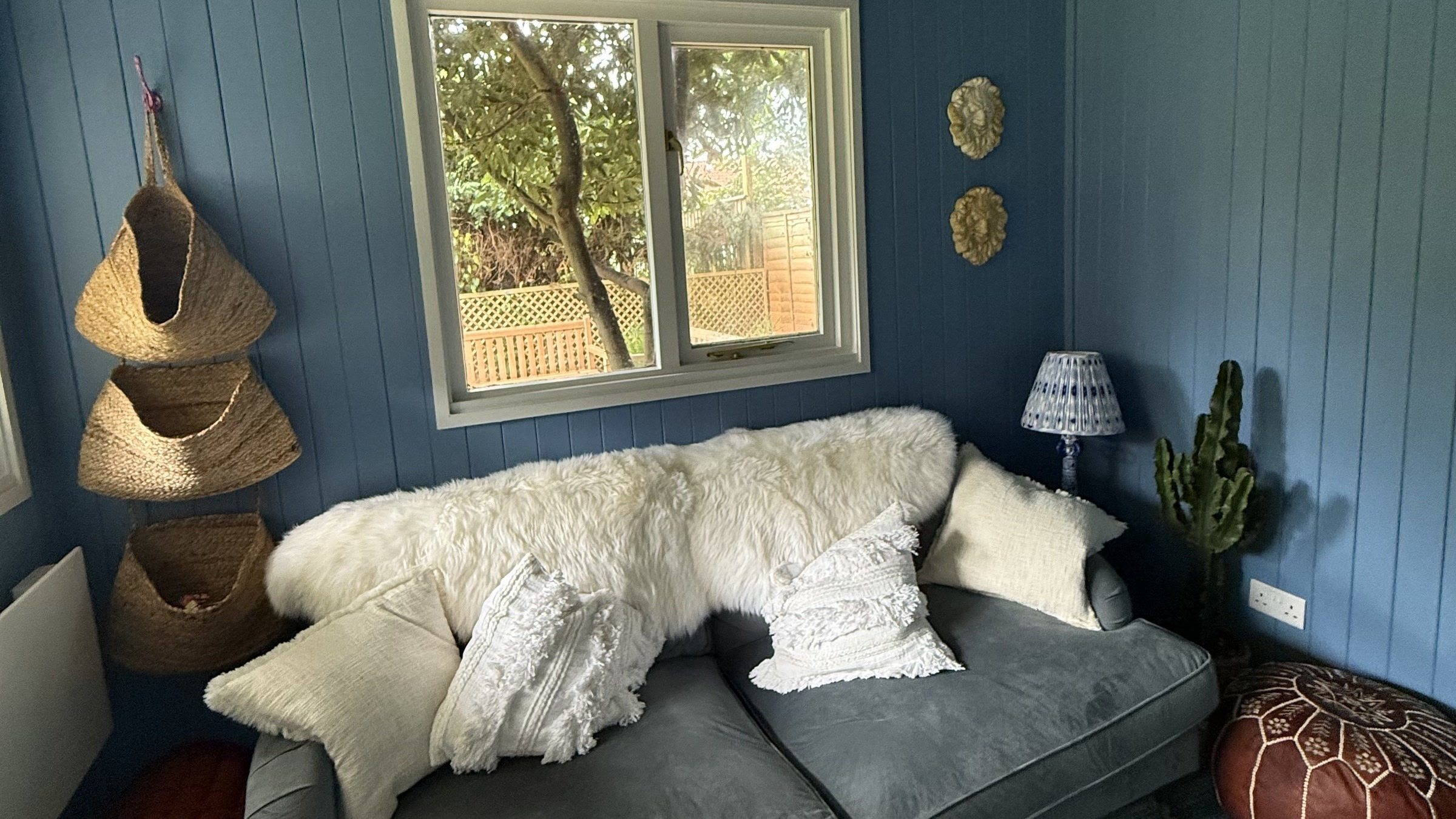 Inside the Cosmic Cabin: a grey sofa under sheepskins below the window, rush baskets on the blue panelled wall