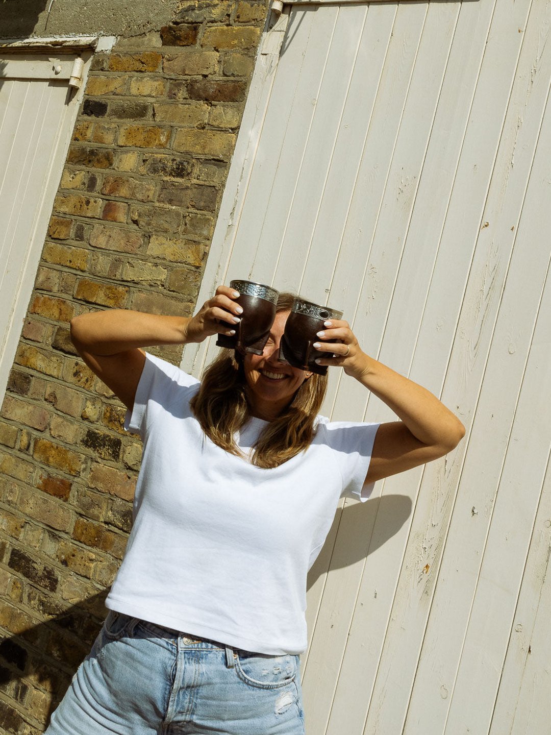 A woman holding two gourds up to her eyes by a white shed door