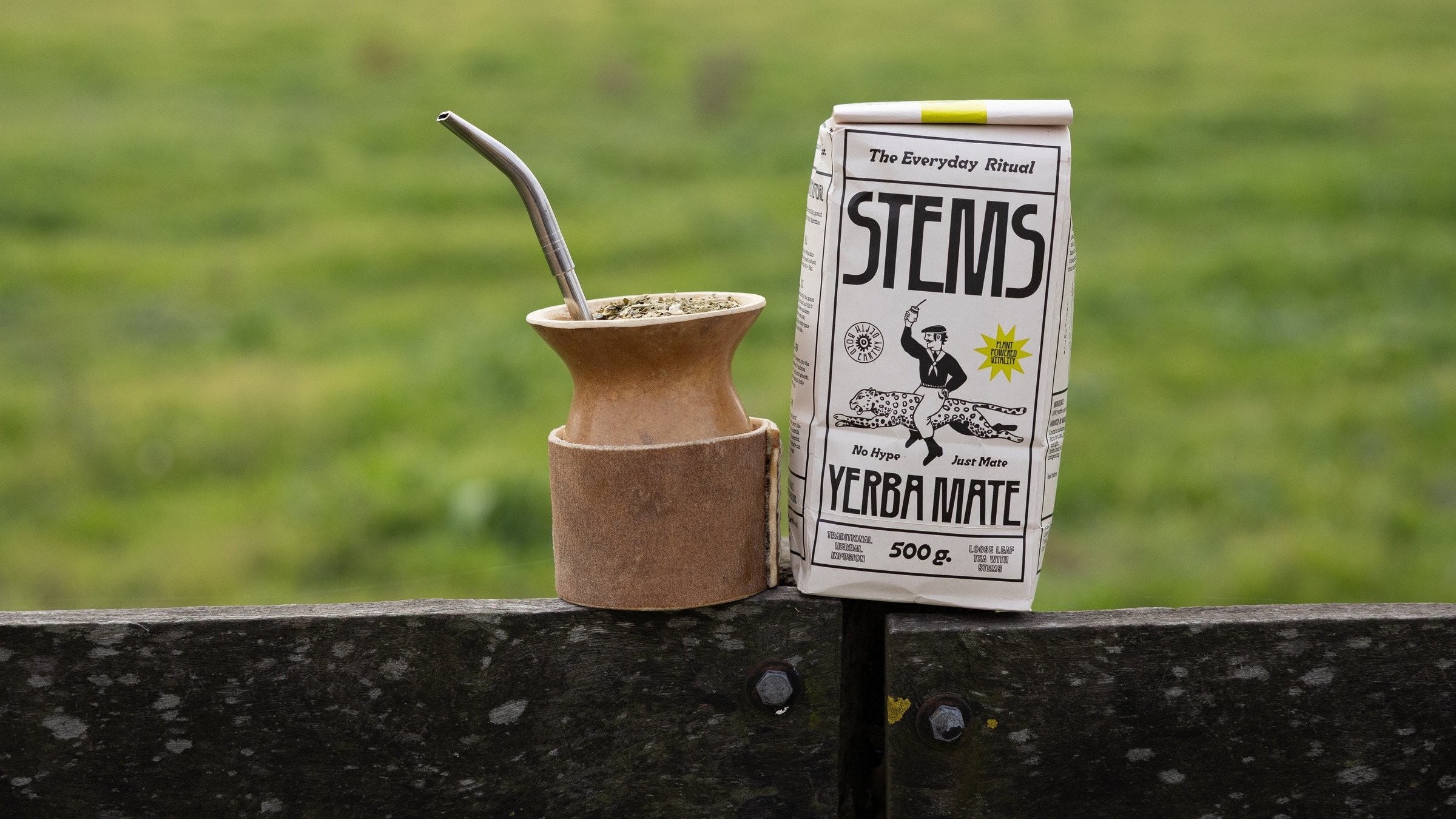 A Stems 500g carton and a mate gourd on a fence rail in front of a green field