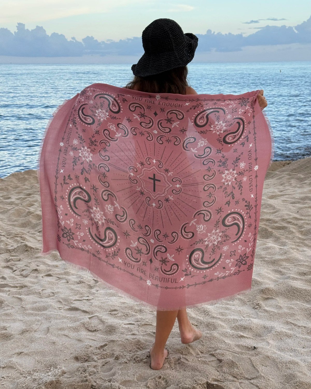 Eli Munroe on a beach, seen from behind, holding the Dusty Rose Wrapped in Truth scarf open wide against the sea