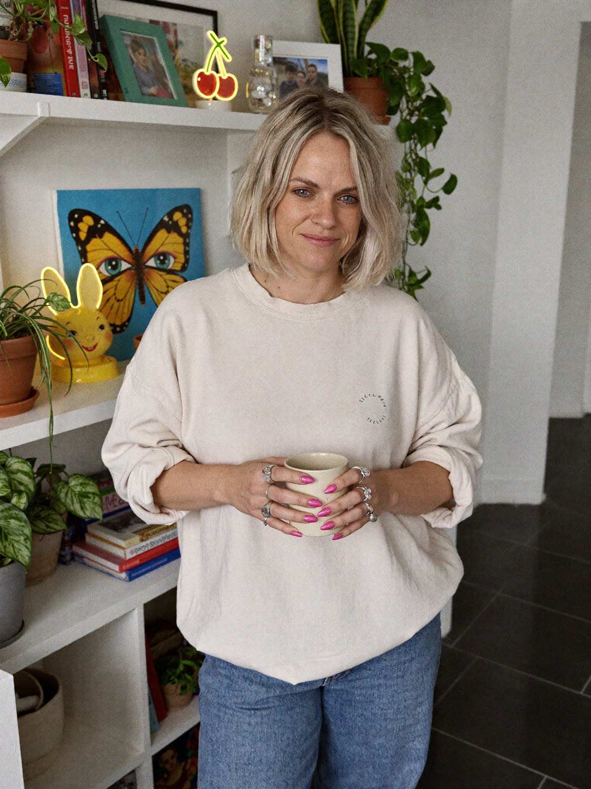 Sophie at home in a cream sweatshirt holding a mug, plant shelf and gallery wall behind her